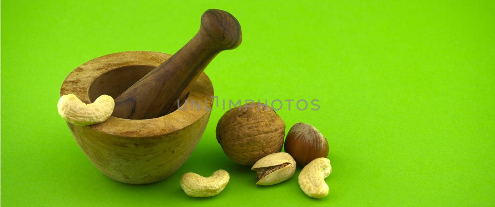 Wooden rustic-style mortar and pestle and various nuts over green background with free space for text. Spices and herb grinder