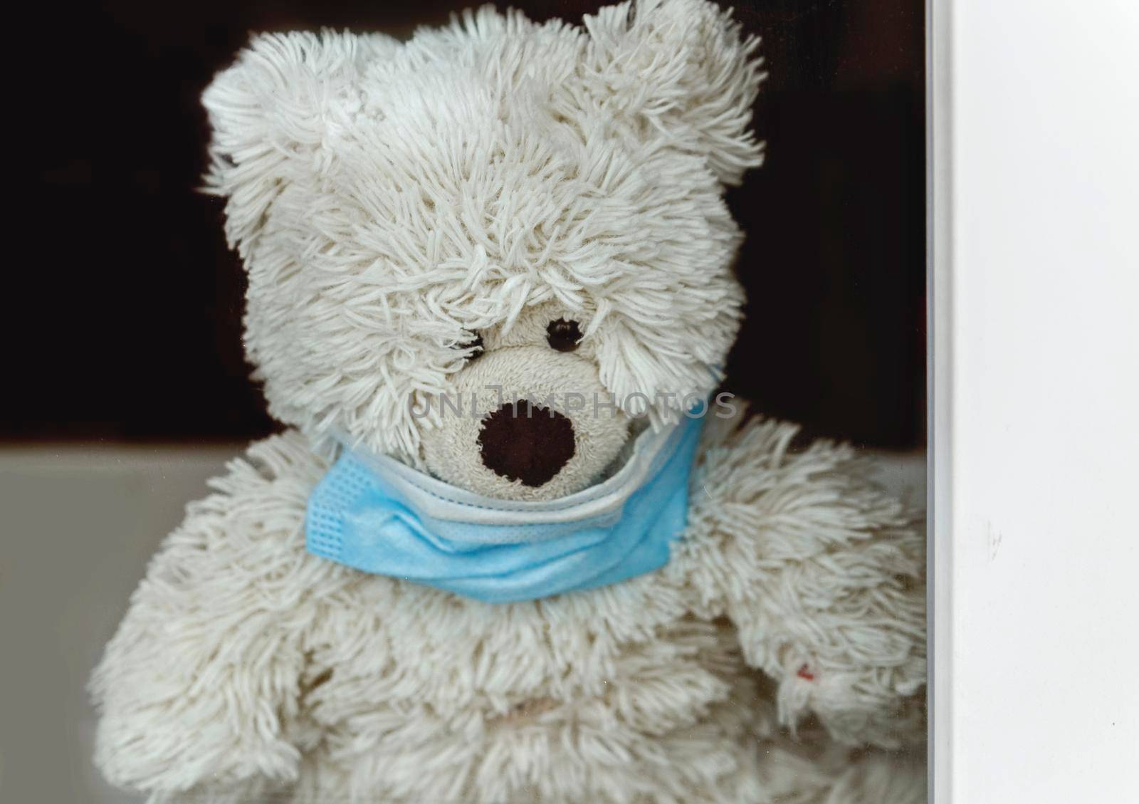 Toy polar bear with medical mask behind window glass. Selective focus