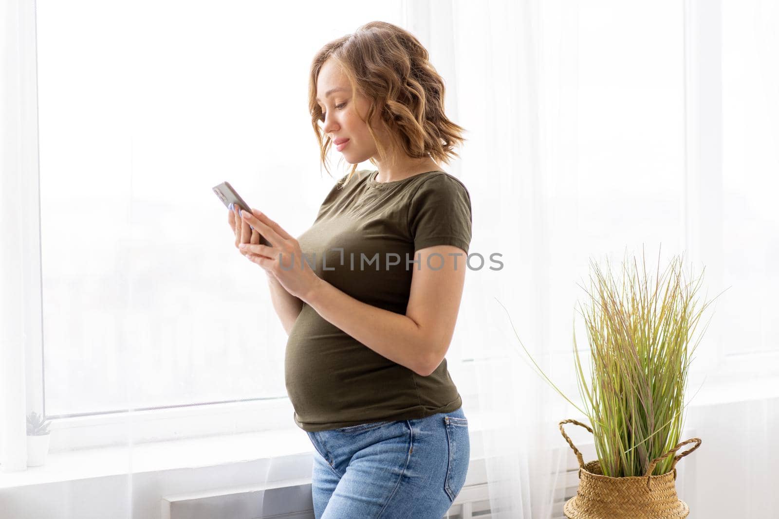 Pregnant woman use smartphone standing near big window Pregnancy Caucasian female with cell phone Modern technology concept