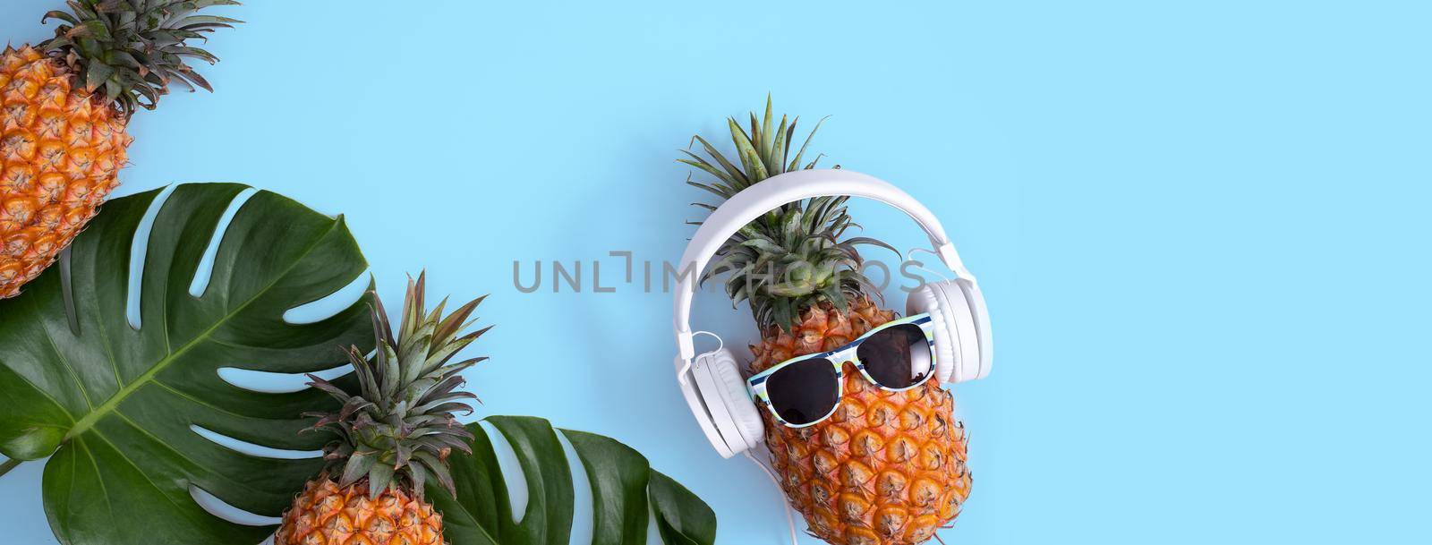 Funny pineapple wearing white headphone, concept of listening music, isolated on blue background with tropical palm leaves, top view, flat lay design.