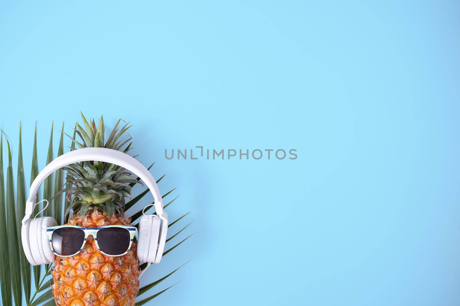 Funny pineapple wearing white headphone, concept of listening music, isolated on blue background with tropical palm leaves, top view, flat lay design.