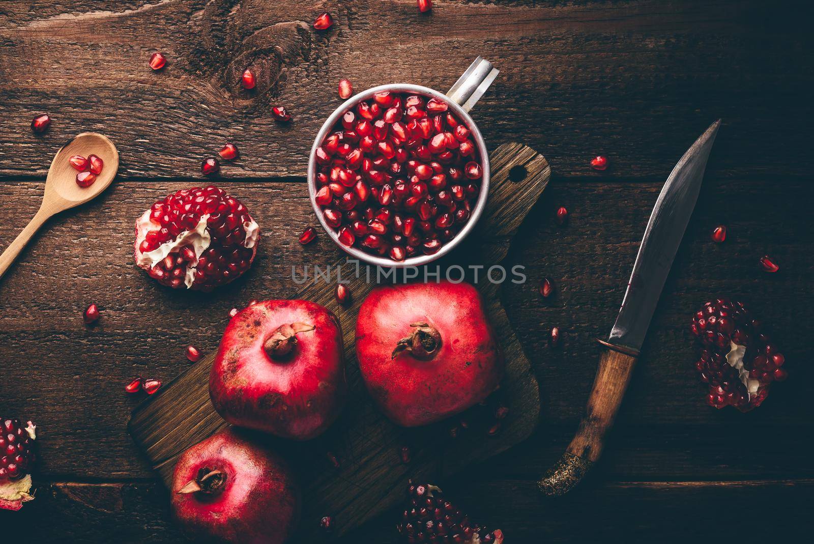 Metal mug full of pomegranate seeds. Whole fruits and pomegranate pieces on rustic wooden table. View from above