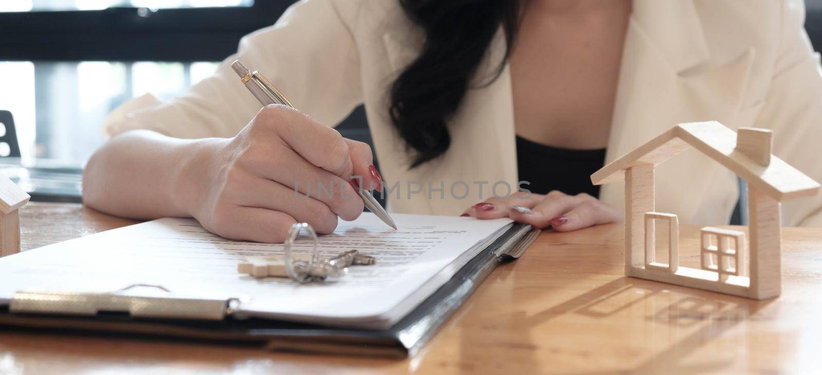 real estate agent assisting client to sign contract paper at desk with house model by wichayada
