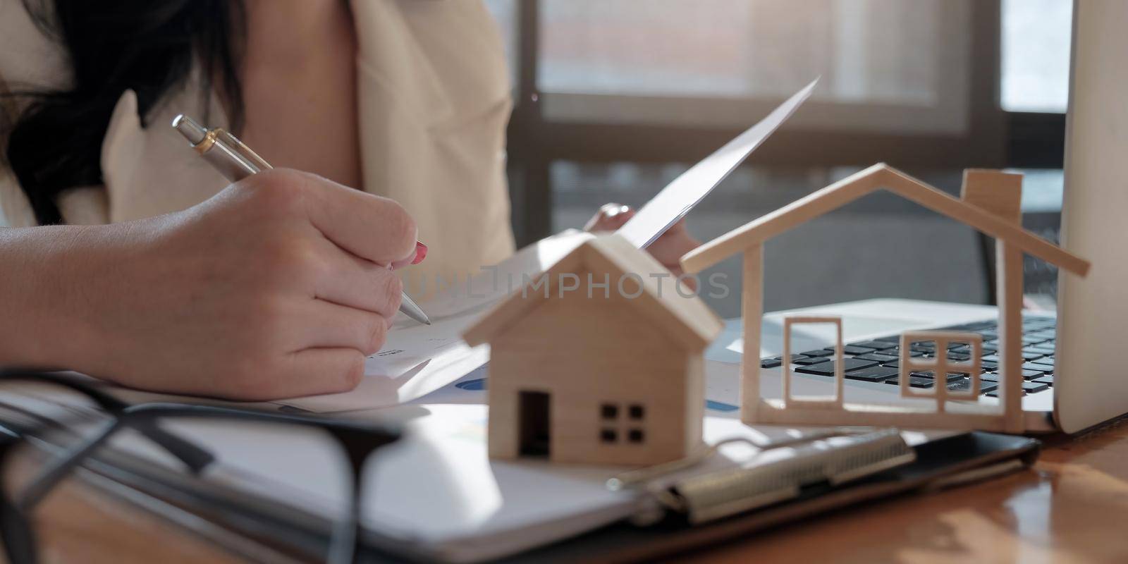 real estate agent assisting client to sign contract paper at desk with house model by wichayada
