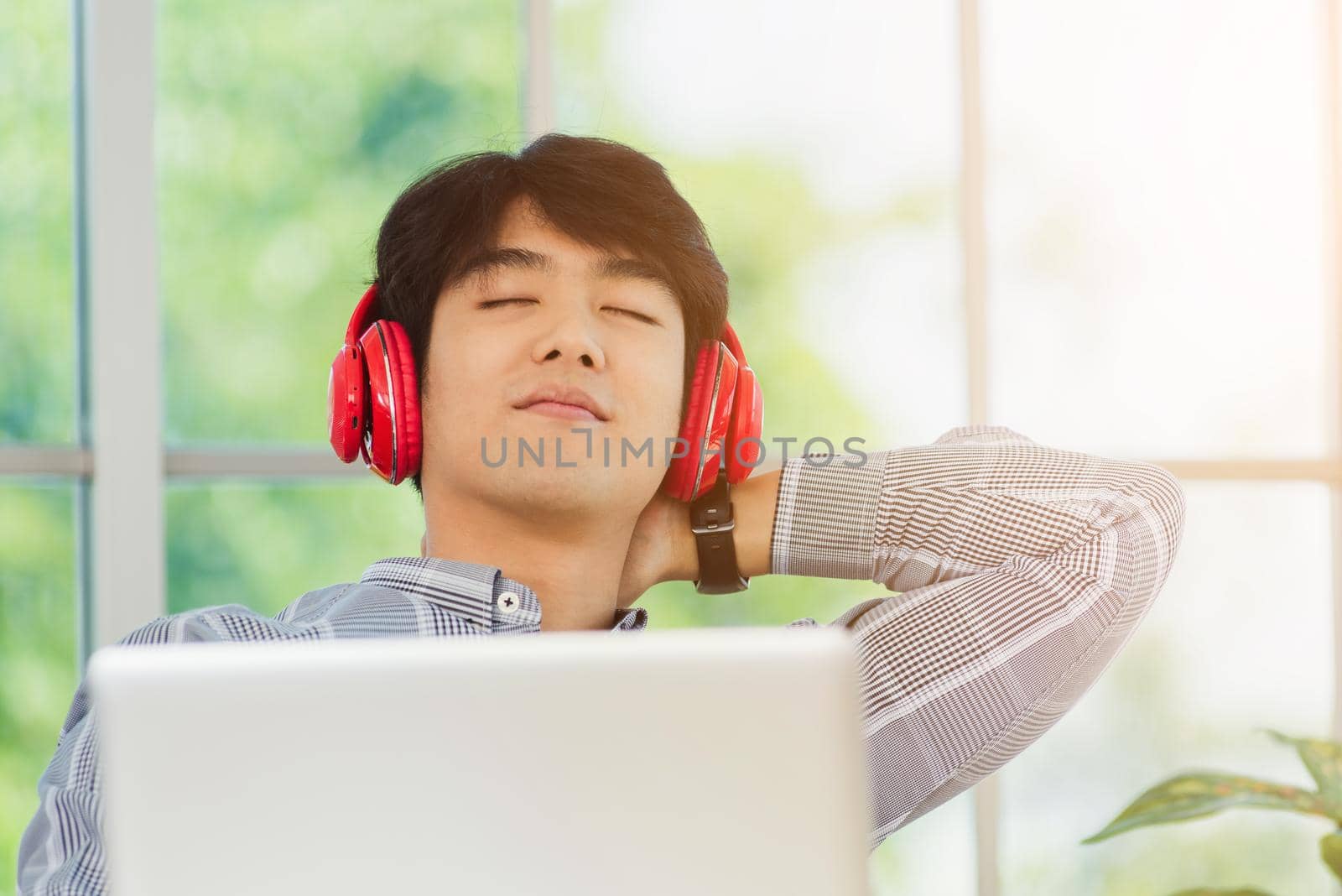 Asian young business man smile listening music in red headphone at home office. Happy businessman sitting on desk and relax wear wireless head phones enjoy listen music and work with laptop computer