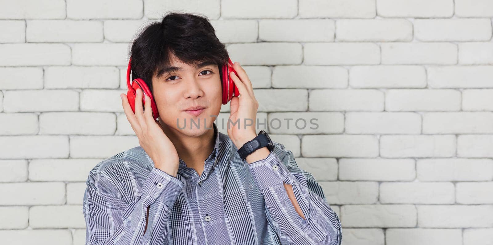 Portrait of Asian handsome young man he smile listening to music in headphones on brick wall background. Happy teenage guy smiling. Happy laughing man standing with red headphones