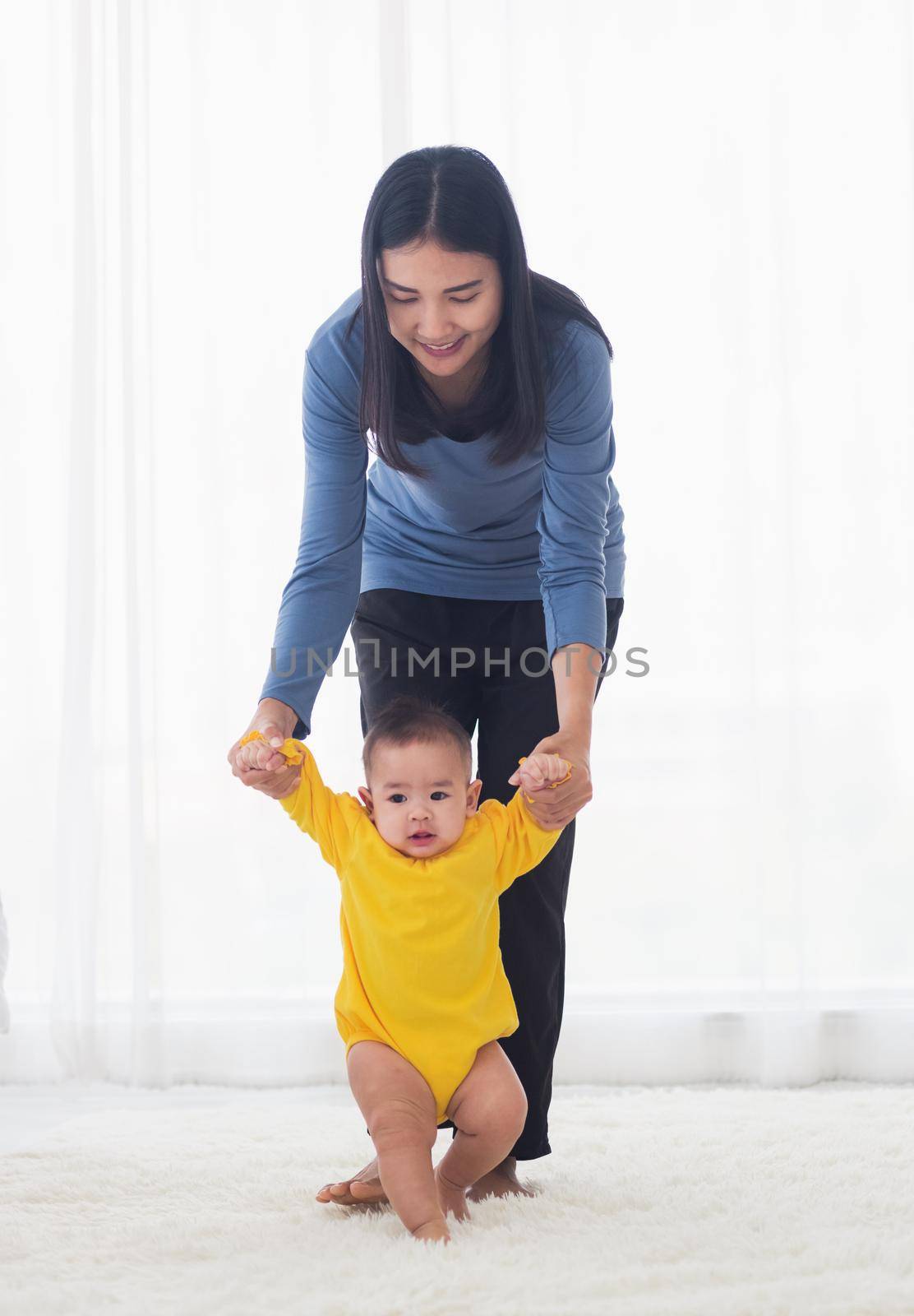 Asian little baby girl taking first steps learning to walk with mom help support the cute unstable walking toddler at home in bedroom. Happy family first steps parenthood concept