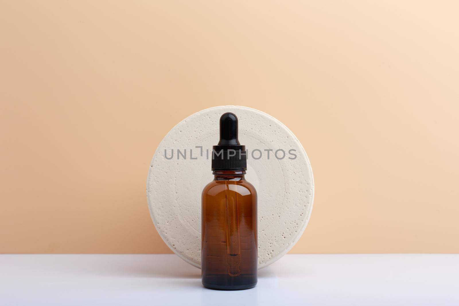 Skin serum in dark brown bottle against round stone circle on white table against beige background with copy space by Senorina_Irina