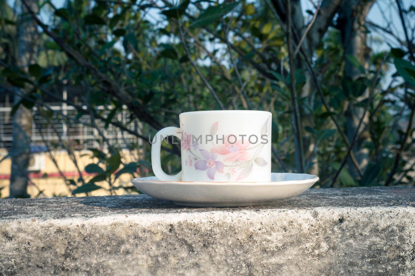 Coffee cup in morning sunlight. Summer fresh cool look. White coffee cup on saucer for hot drink on roof beam of a residential building with bokeh background. by sudiptabhowmick