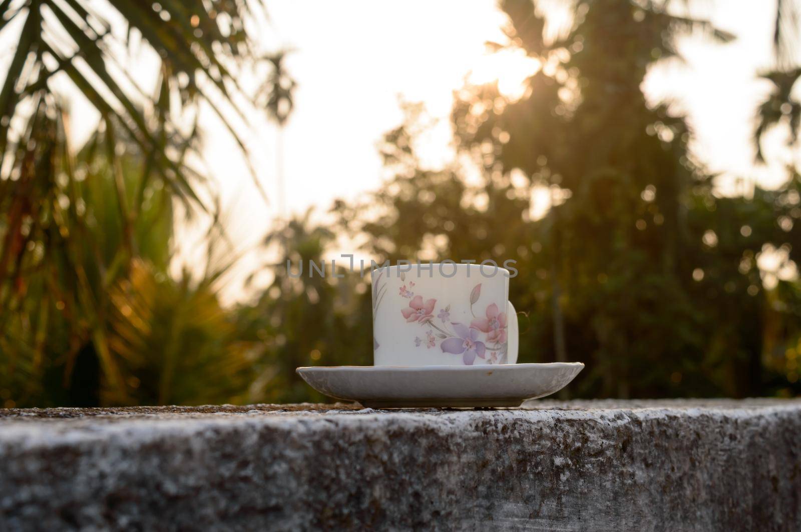 Coffee cup in sunset sunlight. Summer fresh cool look. White coffee cup on saucer for hot drink on roof beam of a residential building with bokeh city in the background. by sudiptabhowmick