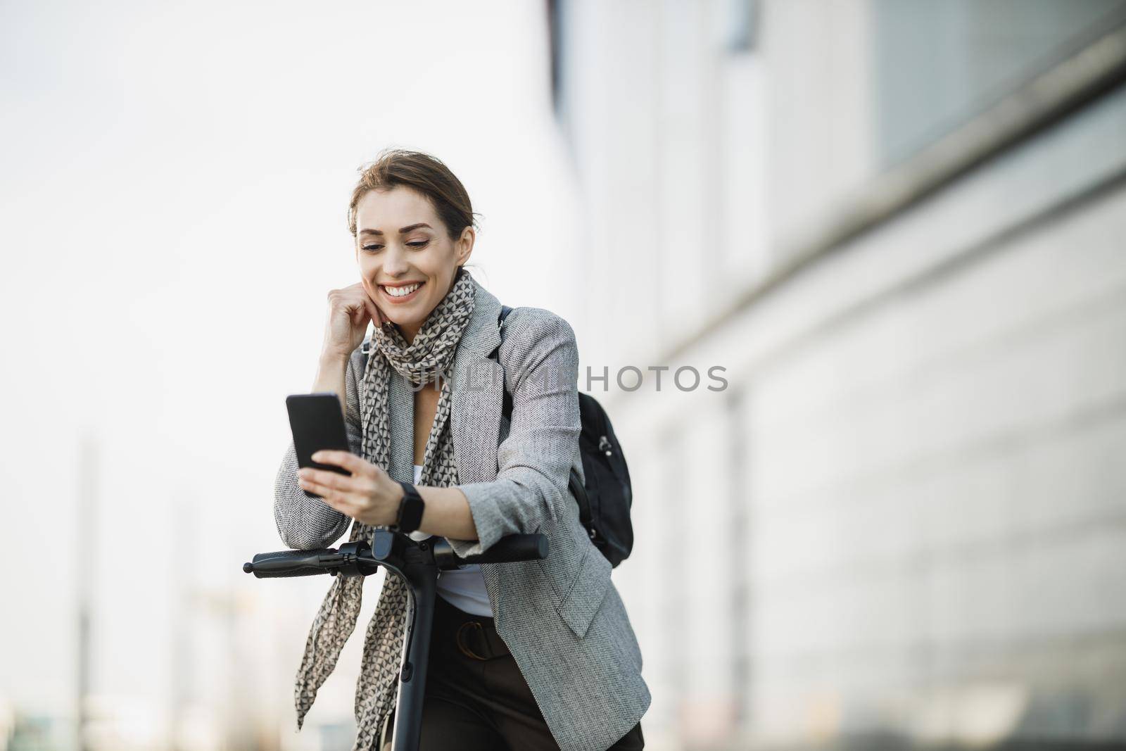 A young businesswoman using a smartphone while traveling with an electric scooter through the city.