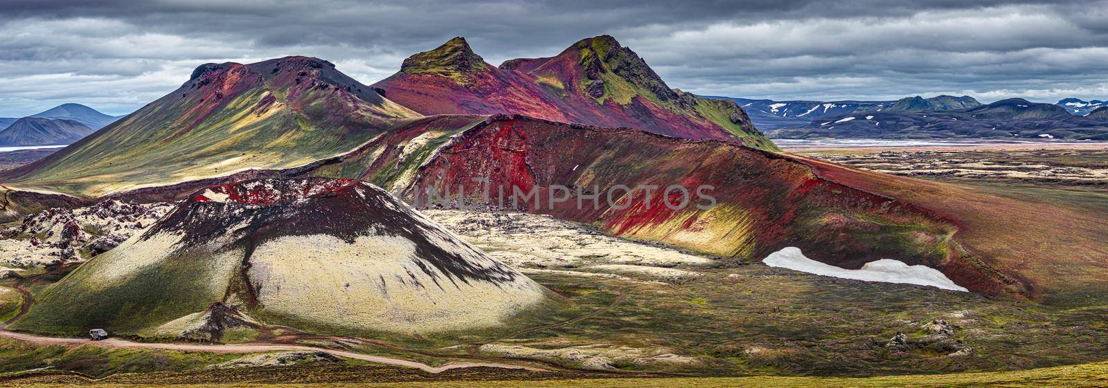Panoramic surreal magic Icelandic landscape of colorful rainbow volcanic Landmannalaugar mountains, red and pink volcanic crater Stutur at famous Laugavegur hiking trail with dramatic sky and a big 4WD car on the road, Iceland
