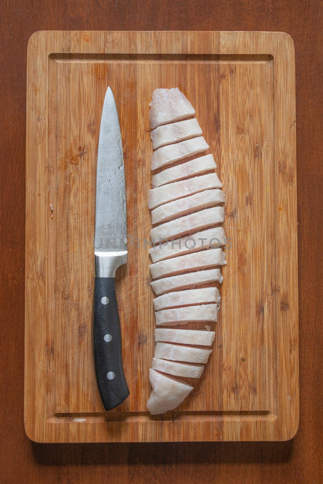 Half frozen fish at a wooden board cut on fine slices by a big knife, details, closeup. Concept home cooking and healthy food.