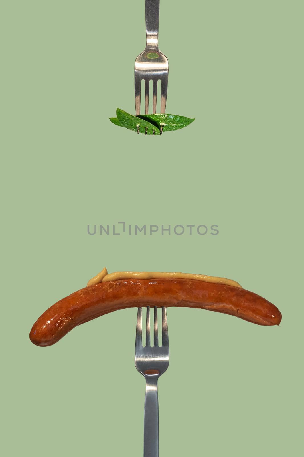 Green fresh leaves picked by a fork for eating as new vegan food and grilled sausage with mustard on another fork at salad color solid background with copy space for text, closeup, details.