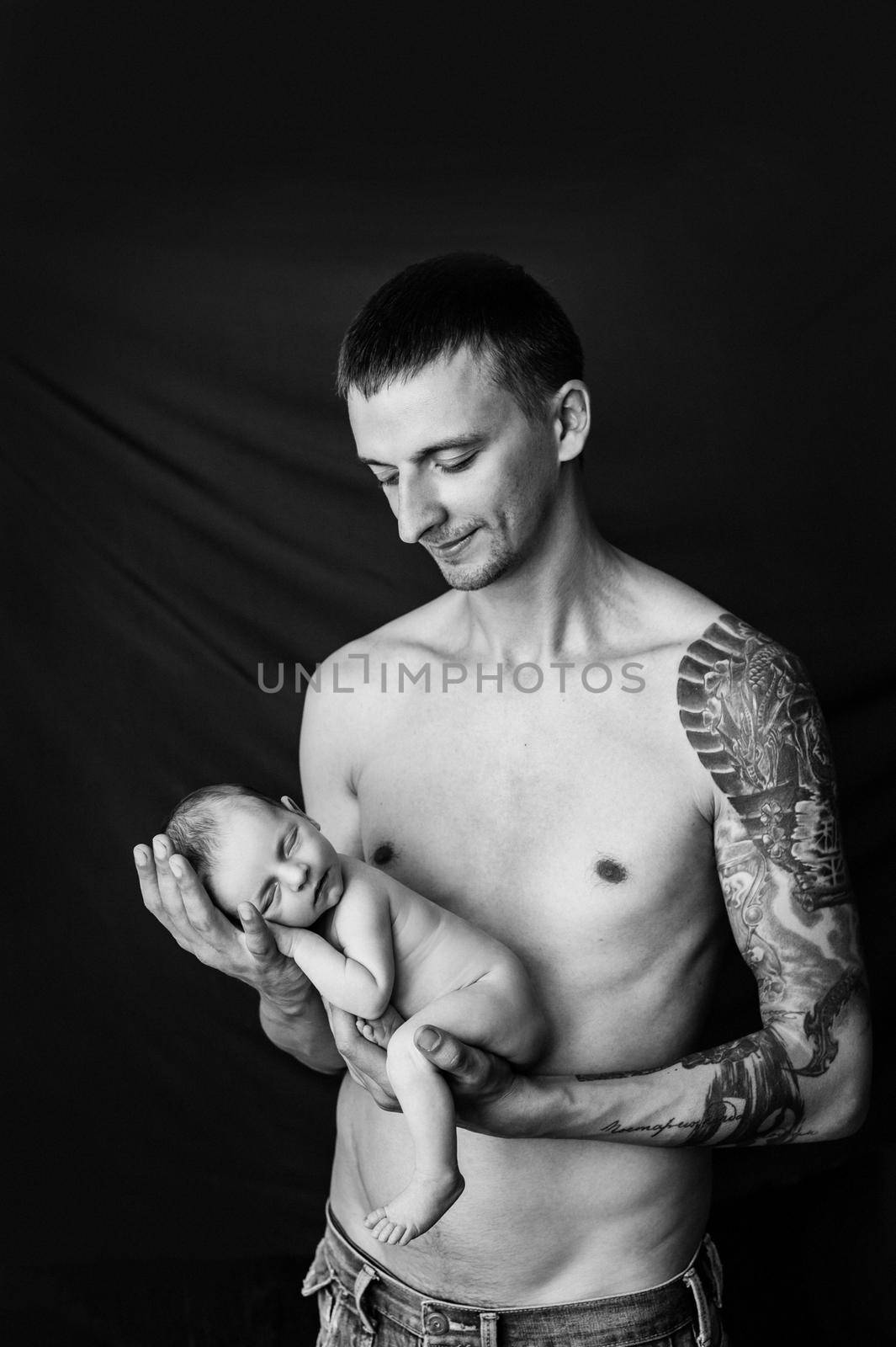 Father with tattoo on his arm is holding his newborn baby on a black and white background. High quality photo