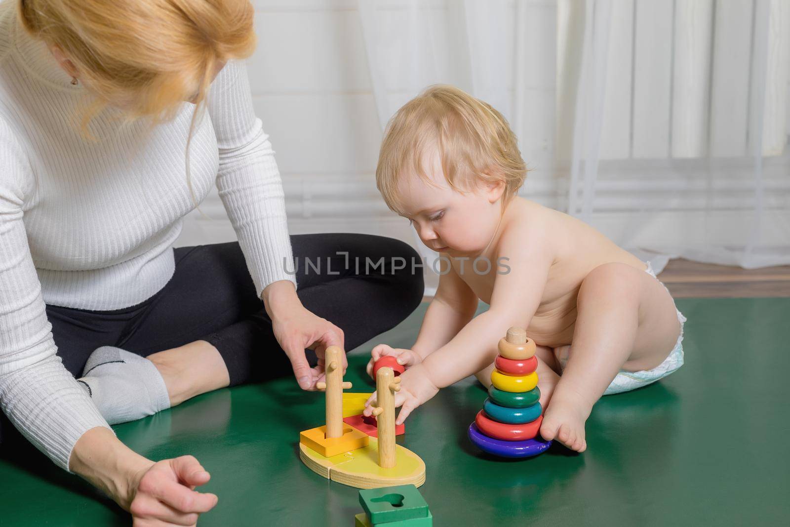 The kid sits on the rug, plays with his mother, collects a pyramid. Close-up.