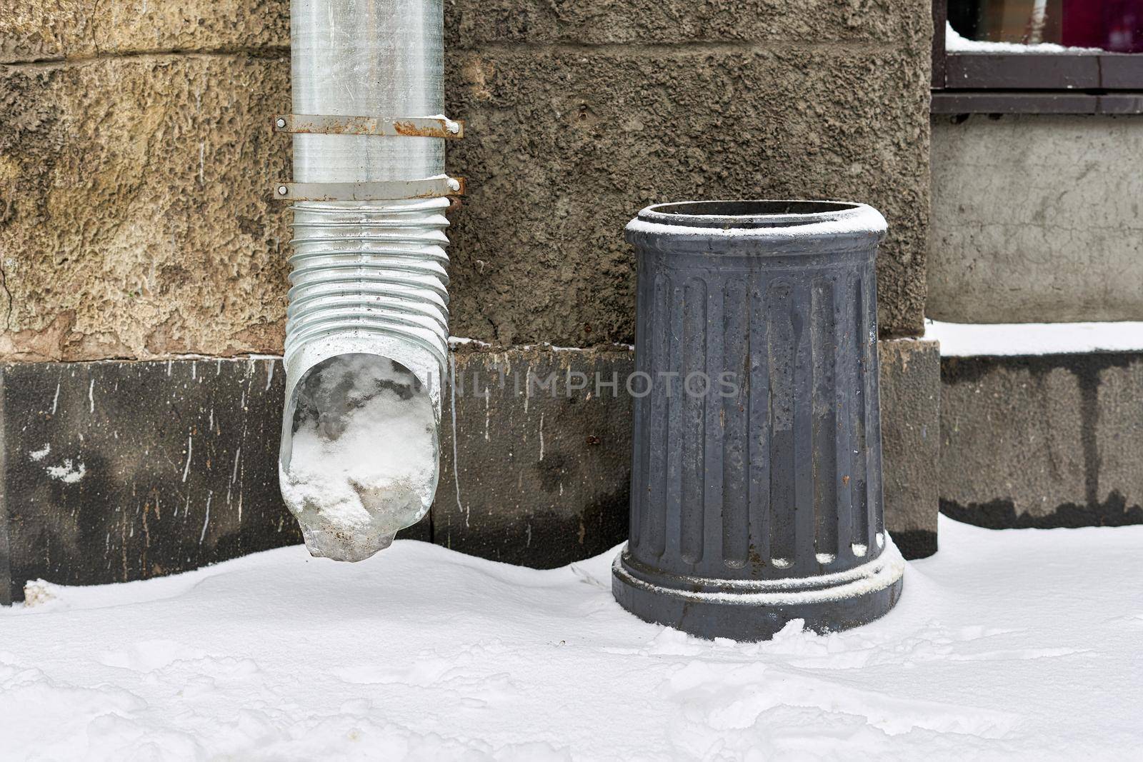 Fragment of a wall with a frozen downpipe and a stone waste basket. Winter street