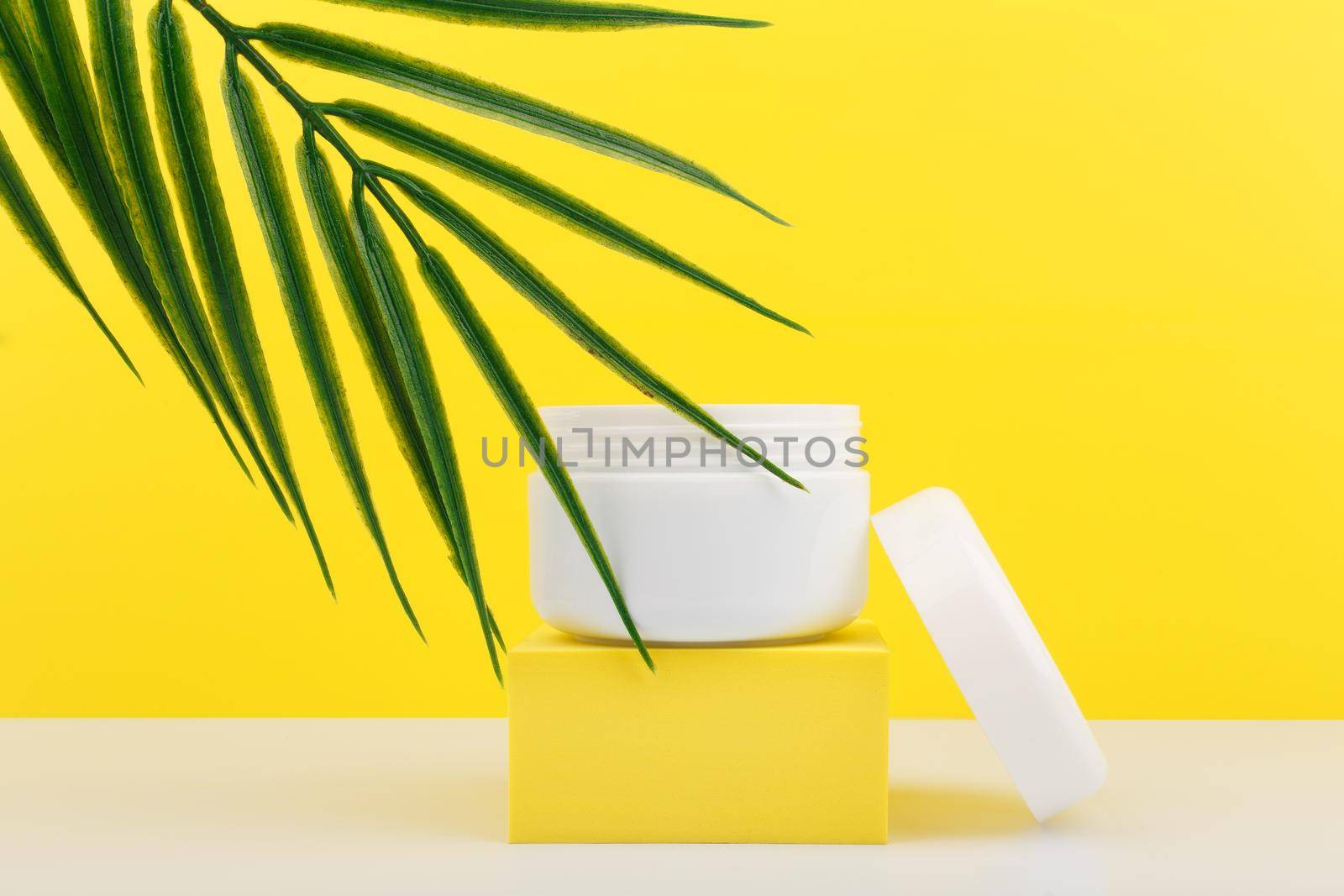 White opened cosmetic jar on yellow podium with palm leaf against bright yellow background. Plastic cosmetic jar for face mask, cream, scrub or balm