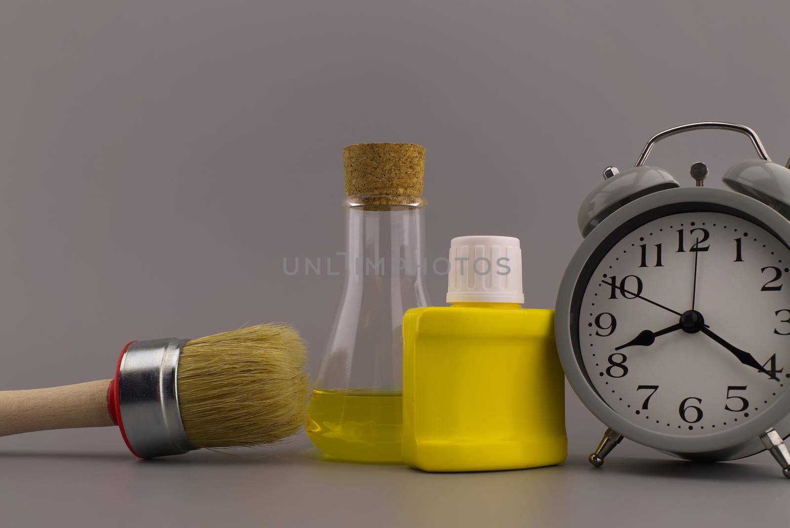 DIY, decorating or renovations concept with a still life of an alarm clock with clean paint brush, yellow pigment container and laboratory flask over a grey background with free space for text