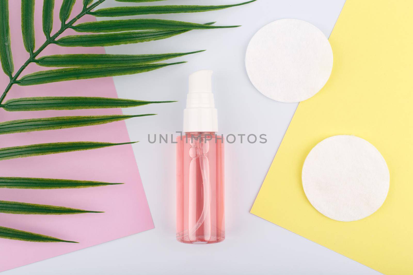 Flat lay with cleansing and soothing foam for make up removing next to cotton pads on pink and yellow pieces of paper and palm leaf. Concept of skin care and make up removing tools