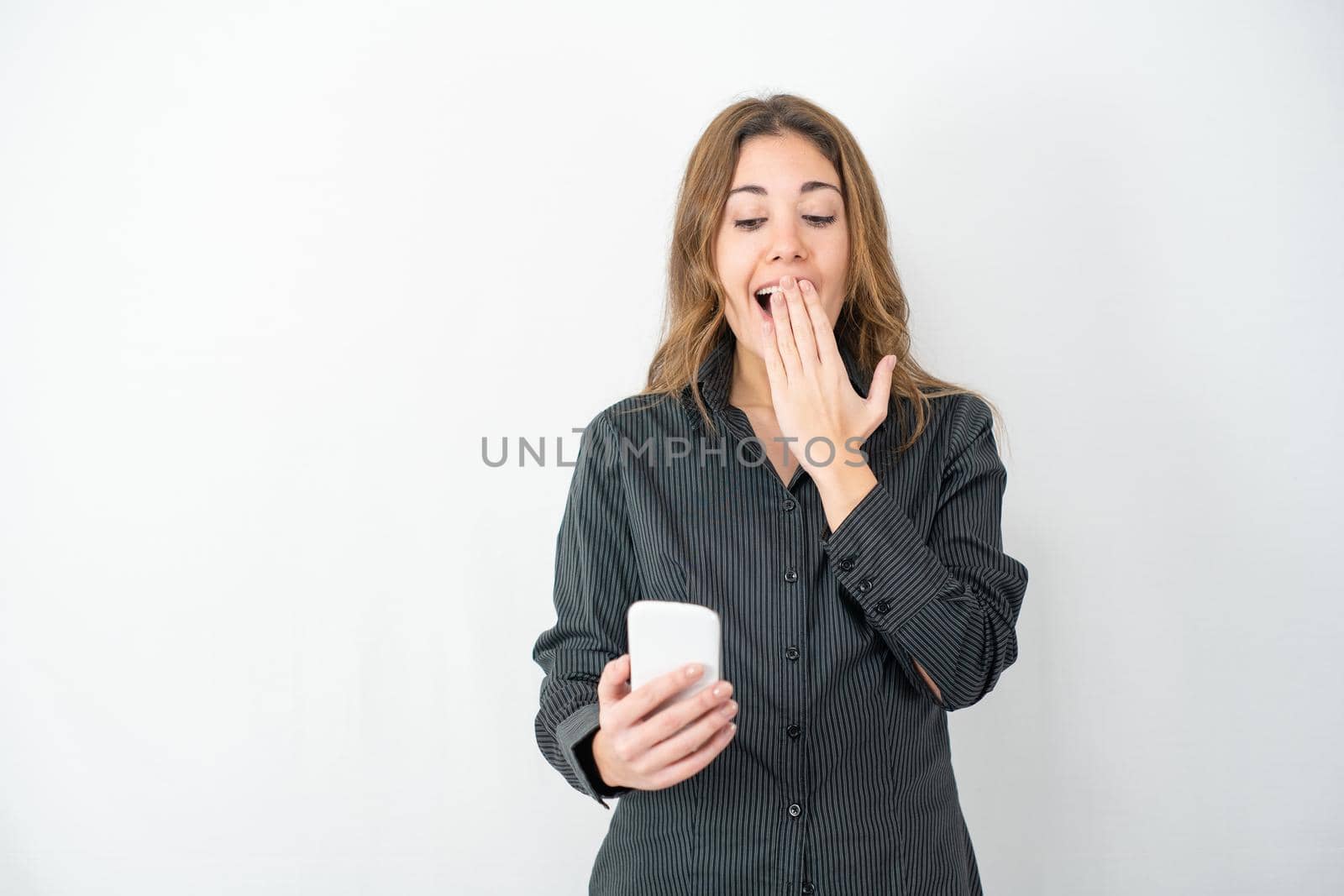 Studio shot of young model woman in grey shirt business clothes isolated on white copy space background surprised looking at smartphone with wow expression hand on mouth. Concept of new technologies