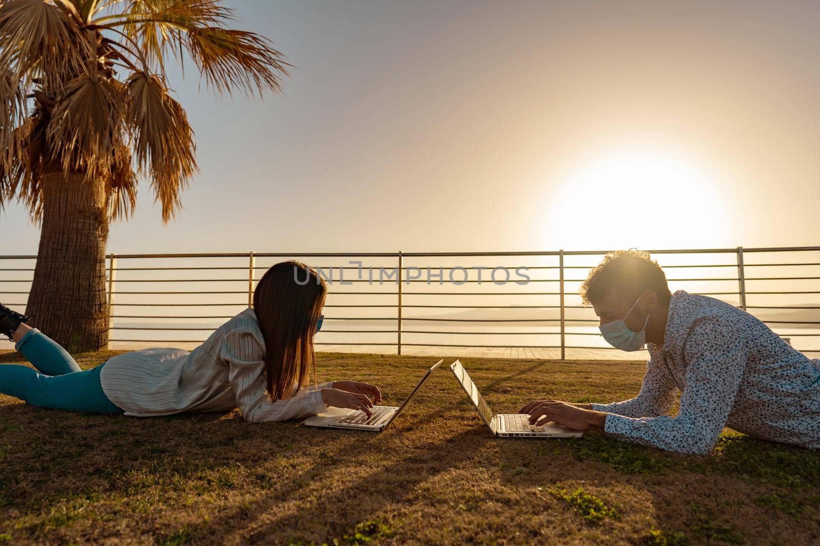 Two students lying on a meadow at sunset learning lesson using laptop computer in the city park of tropical ocean sea resort. Young self entrepreneur couple working for themselves by mobile technology by robbyfontanesi