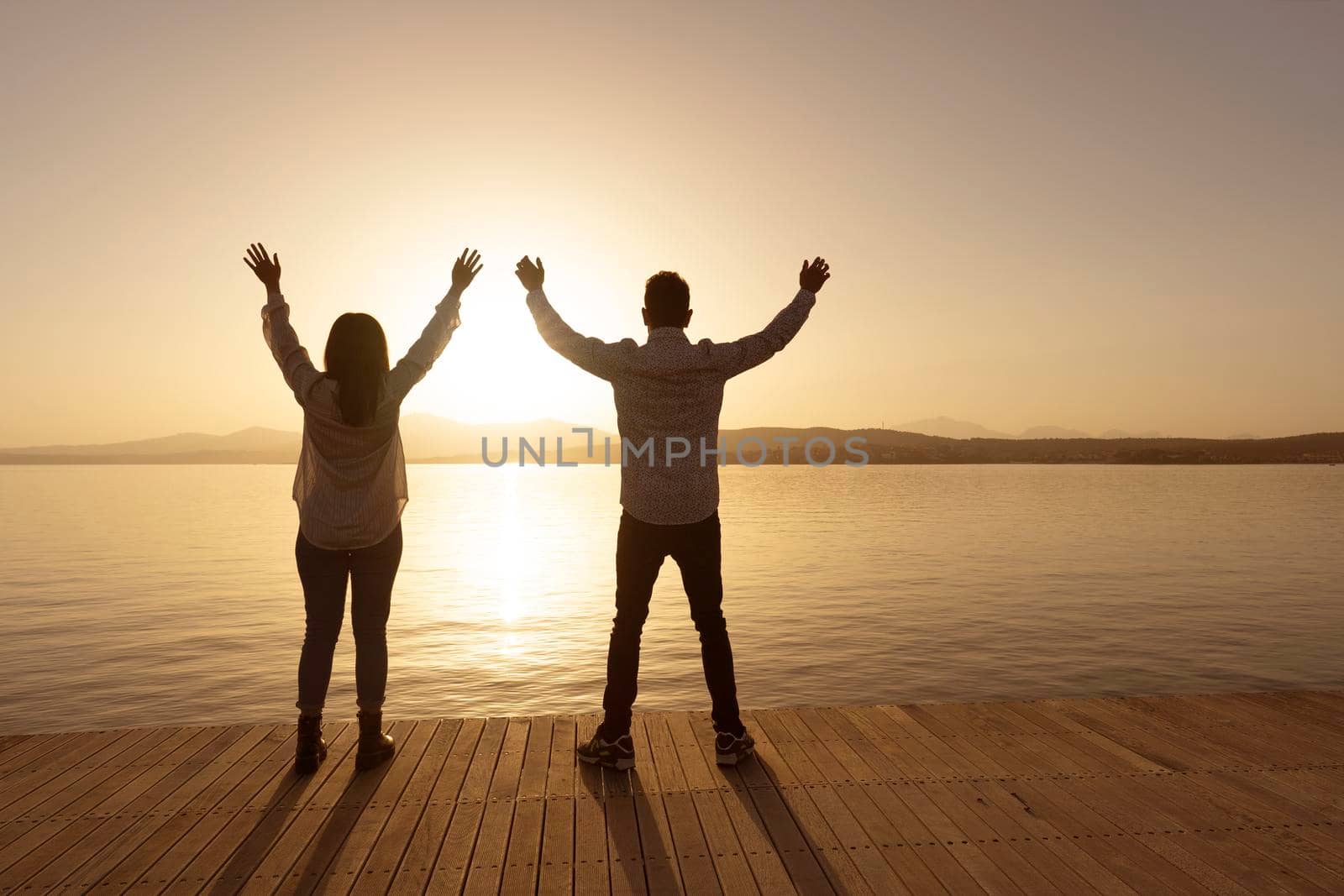 Confidence in the future: view from back silhouette of young couple with open arms looking at forward horizon with rising bright sun on sea ocean water with light reflections. Spiritual moment concept by robbyfontanesi