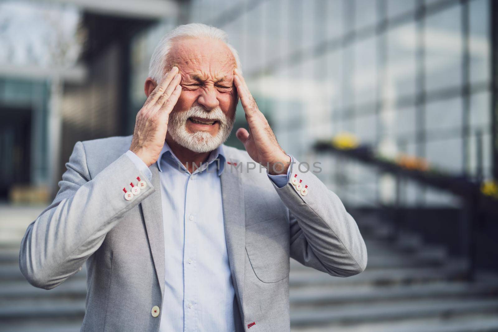 Senior businessman is tired of work. He is standing in front of company building and having headache.