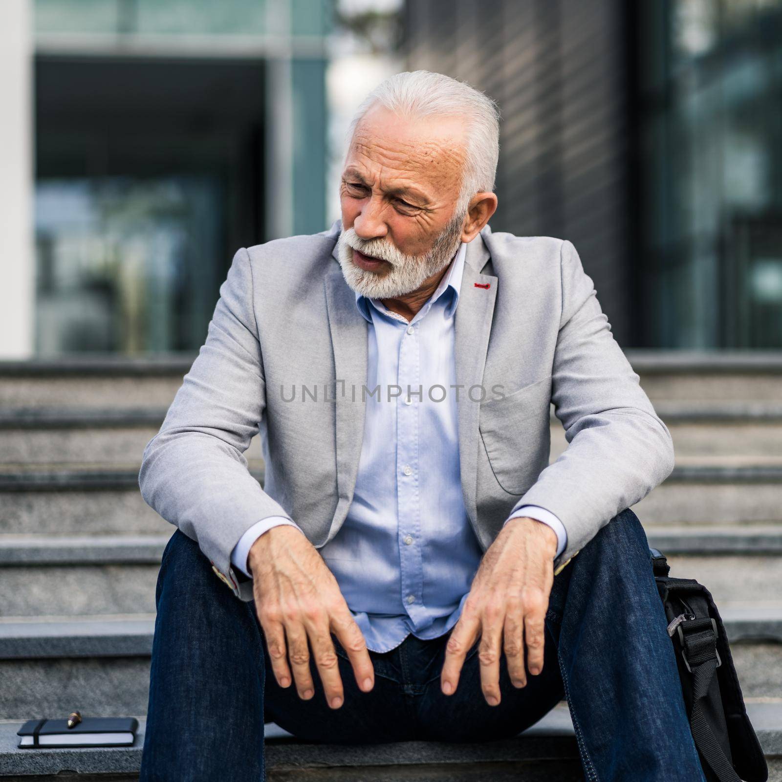 Senior businessman is tired of work. He is sitting on stairs in front of company building.