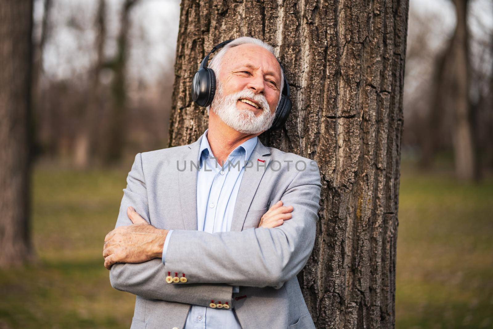 Outdoor portrait of senior businessman relaxing in park. He is listening music on headphones.