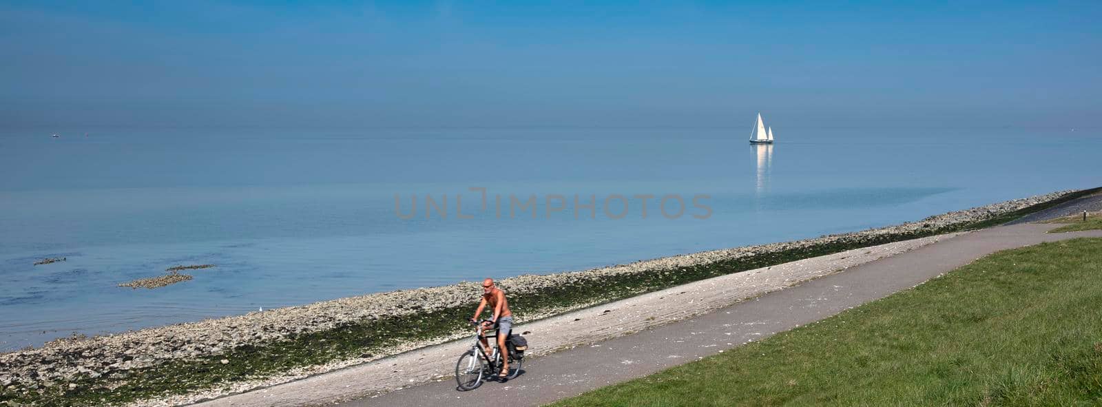 man on bicycle track and lonely sailing boat on vast empty blue lake in zeeland by ahavelaar