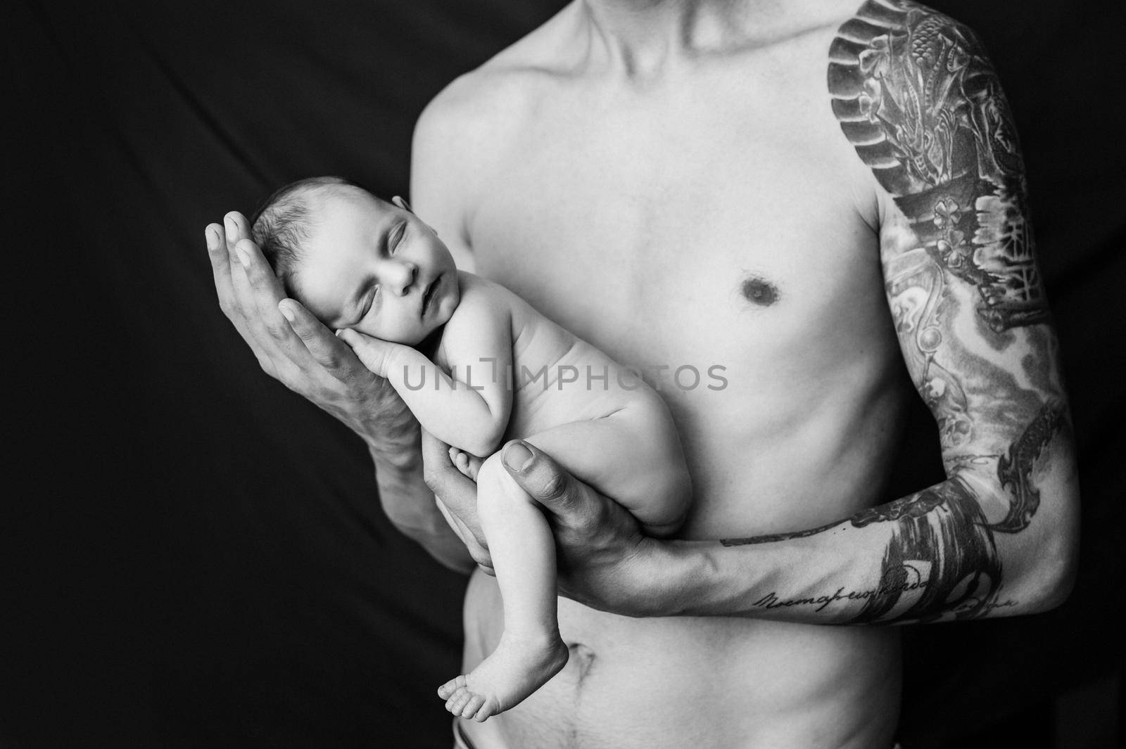 Father with tattoo on his arm is holding his newborn baby on a black and white background. High quality photo