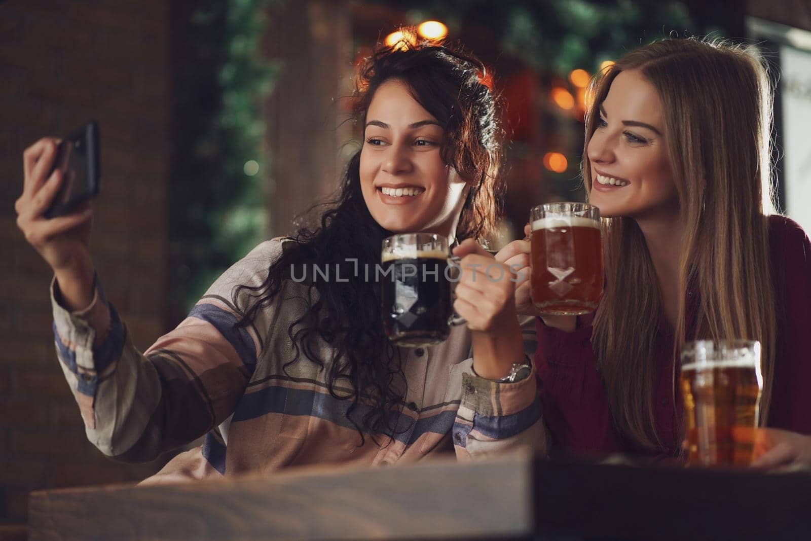 Two happy girlfriends are having fun time in pub. They are drinking beer and taking selfie. Friendship concept.