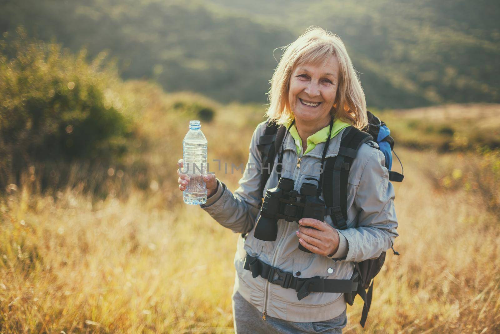 Senior woman is hiking in mountain. Active retirement. She is drinking water.