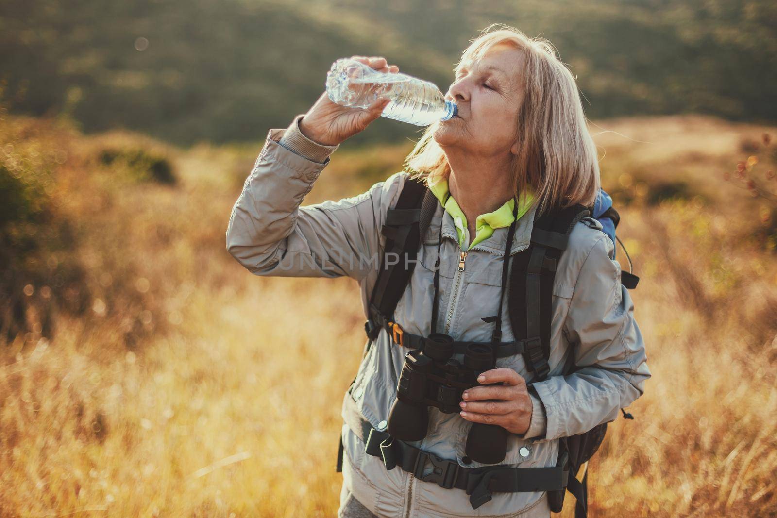Senior woman is hiking in mountain. Active retirement. She is drinking water.
