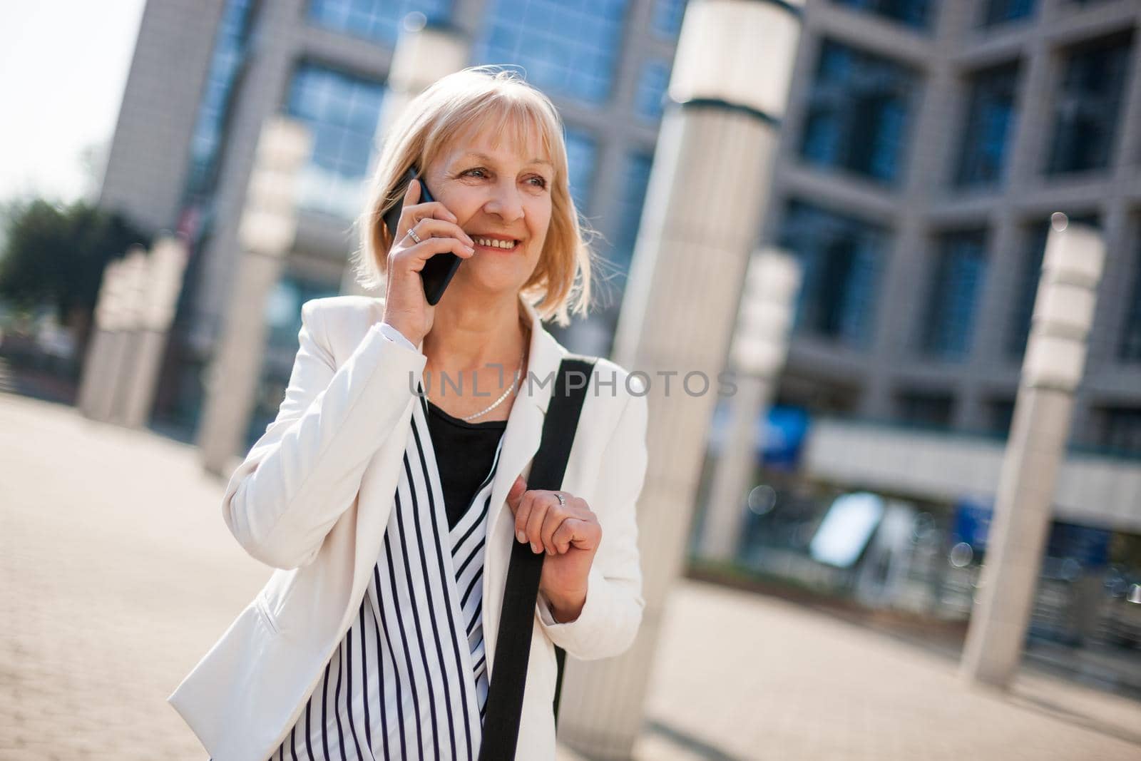 Happy senior businesswoman is talking on phone outside the company building.
