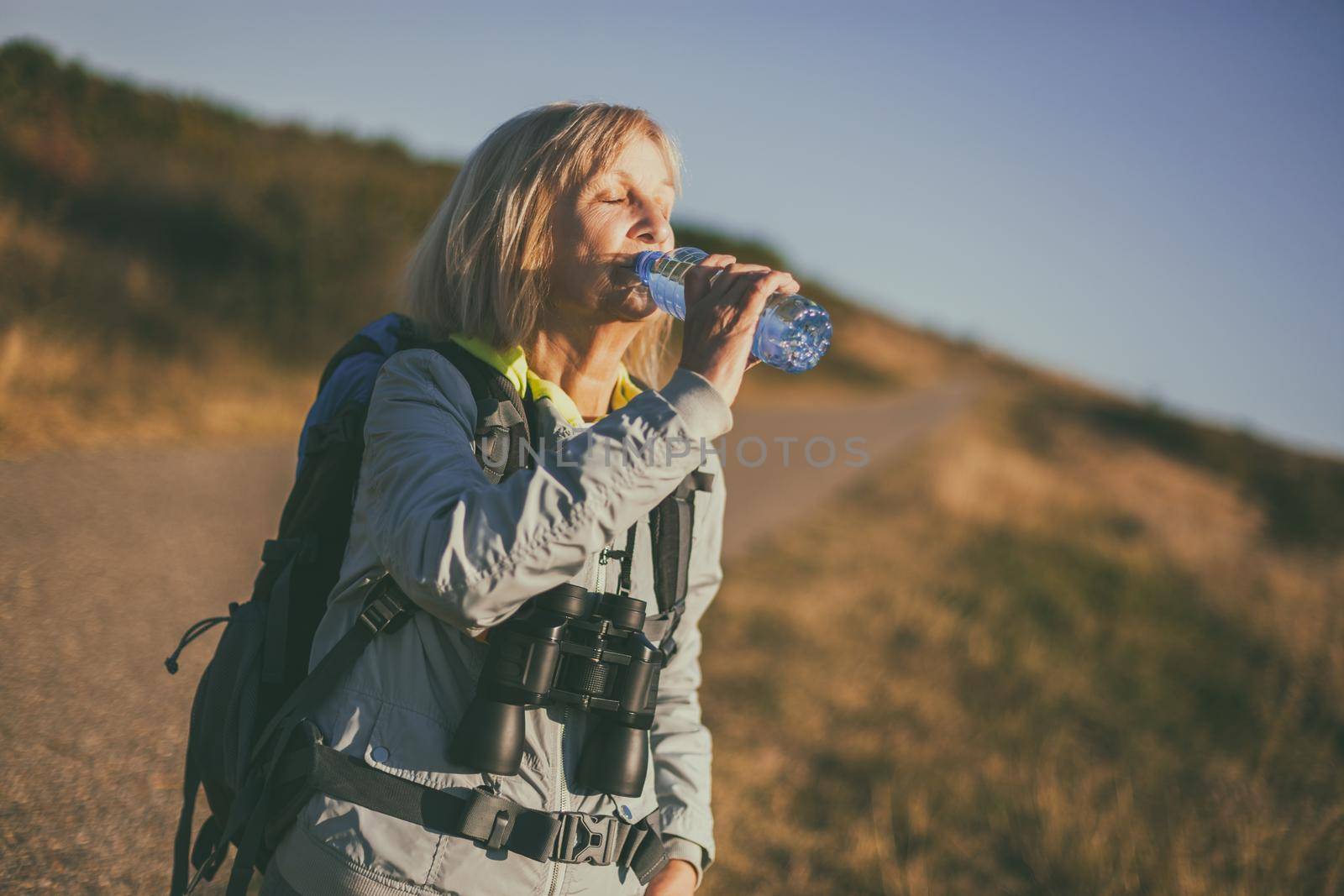 Senior woman is hiking in mountain. Active retirement. She is drinking water.