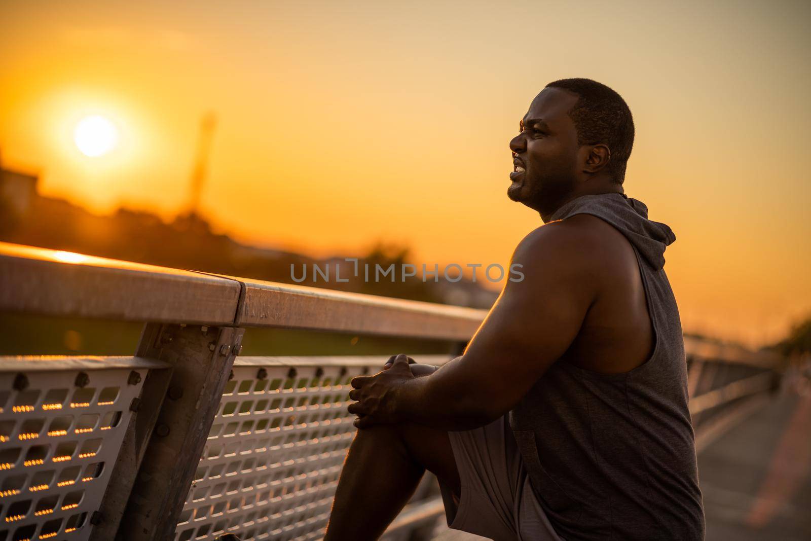 Young african-american man is having pain in knee while exercising in sunset on the bridge in the city.