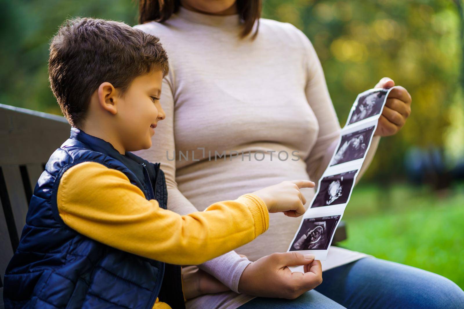 Happy boy and his pregnant mother are enjoying autumn in park. They are looking at x-ray images of baby.