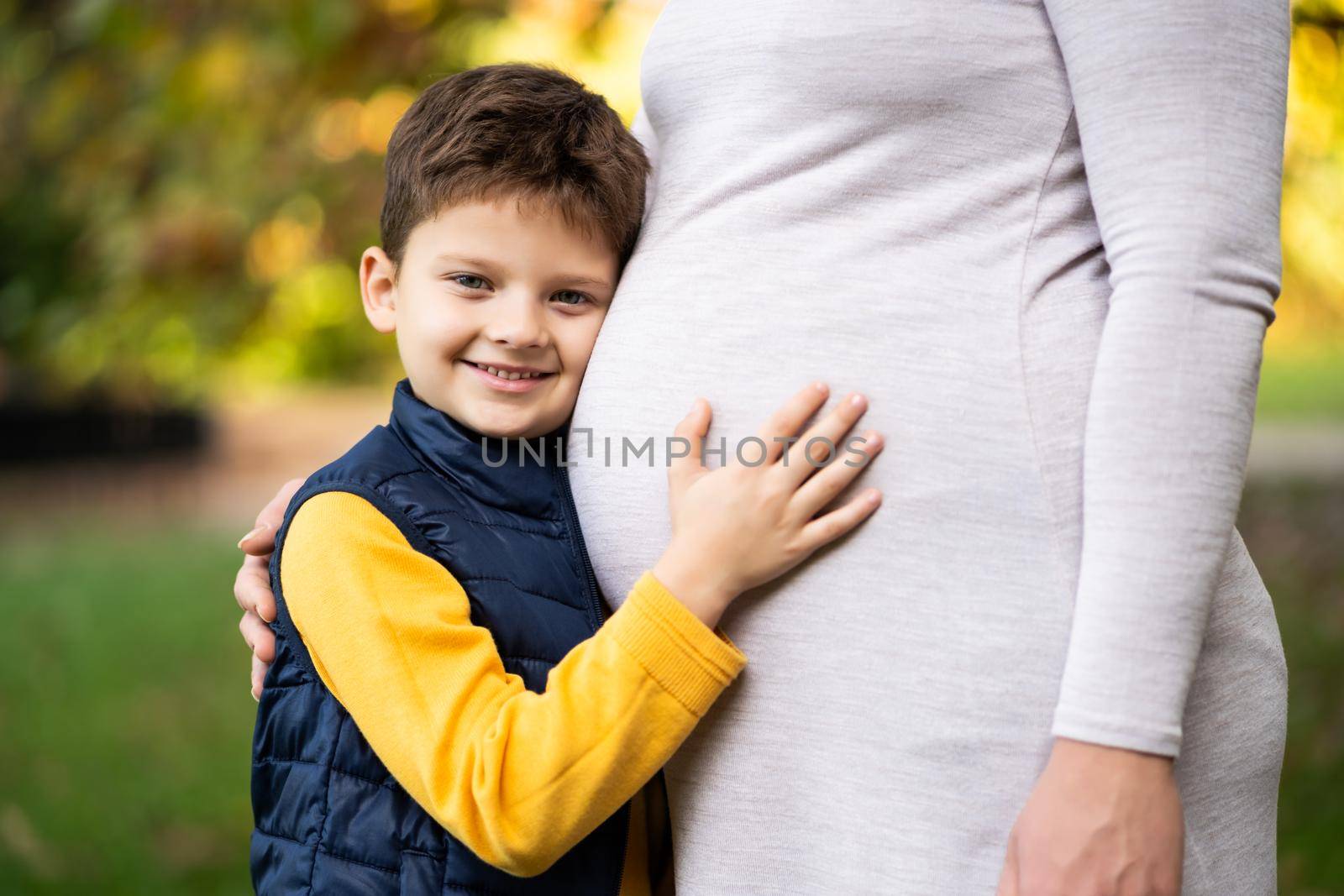 Boy is leaning on stomach of his pregnant mom in park in autumn. Family relaxing time in nature.