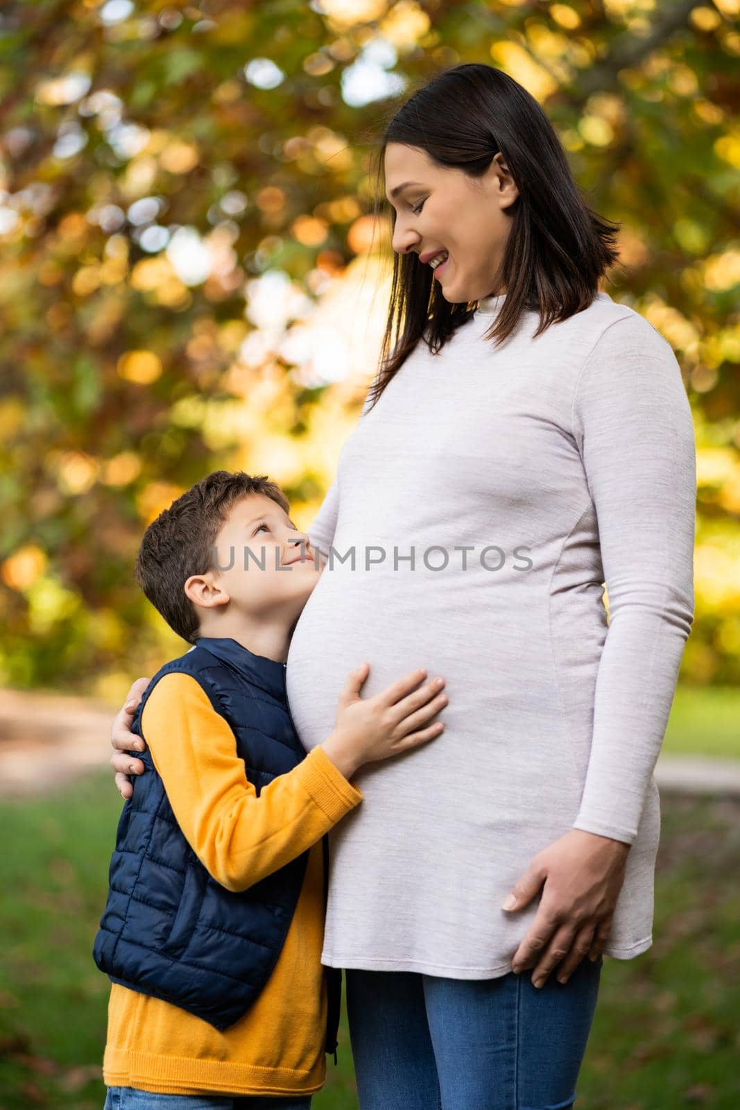 Happy boy and his pregnant mother are embracing in park in autumn. Family relaxing time in nature.