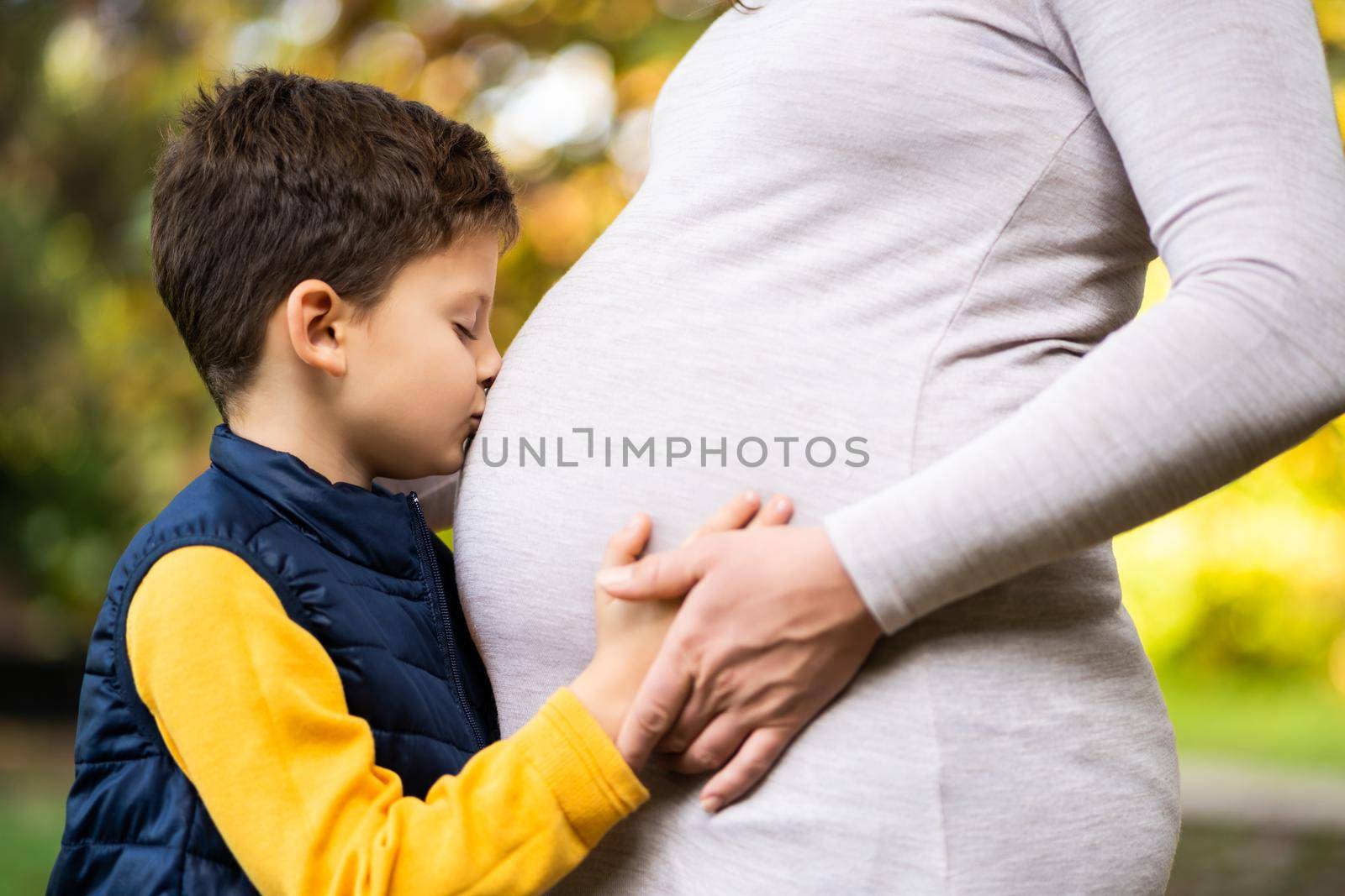 Boy is kissing stomach of his pregnant mom in park in autumn. Family relaxing time in nature.