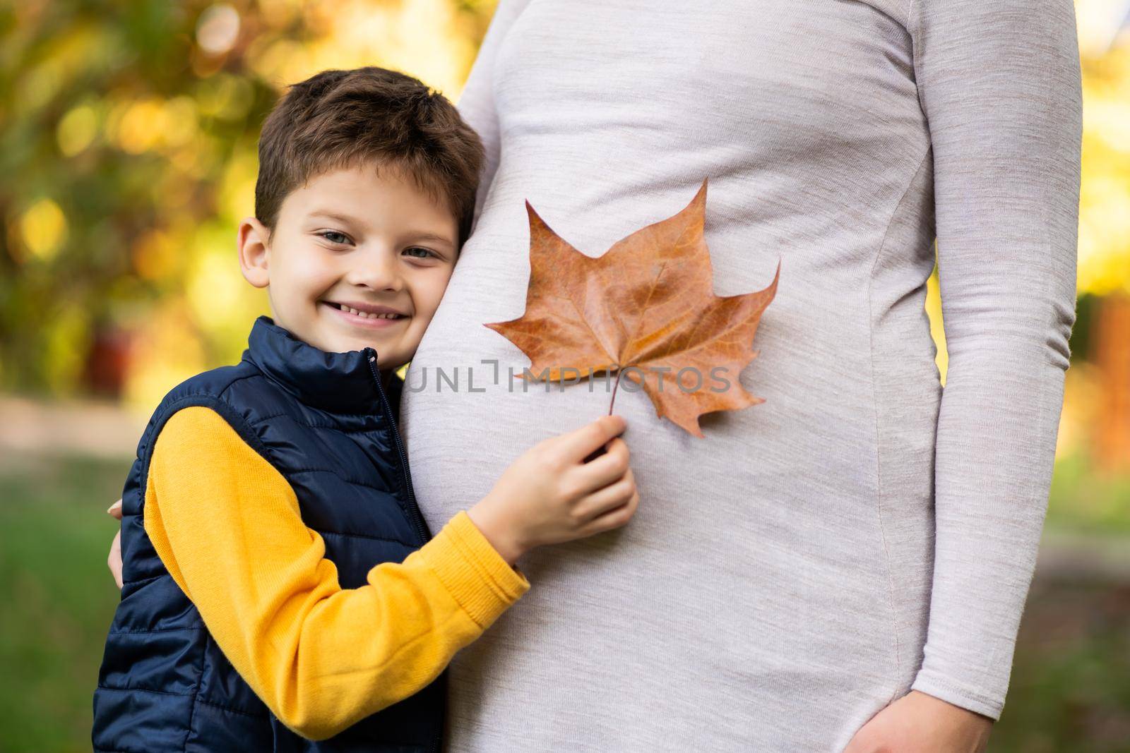 Happy boy and his pregnant mother are embracing in park in autumn. Family relaxing time in nature.