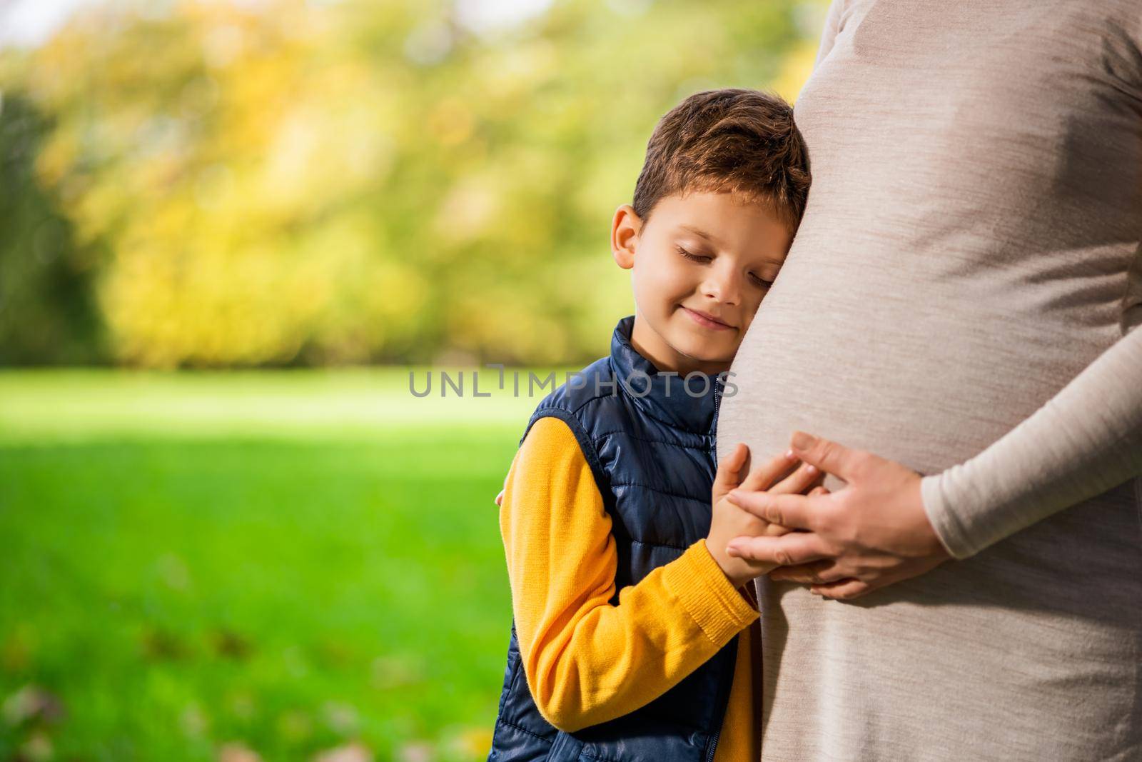 Boy is leaning on stomach of his pregnant mom in park in autumn. Family relaxing time in nature.