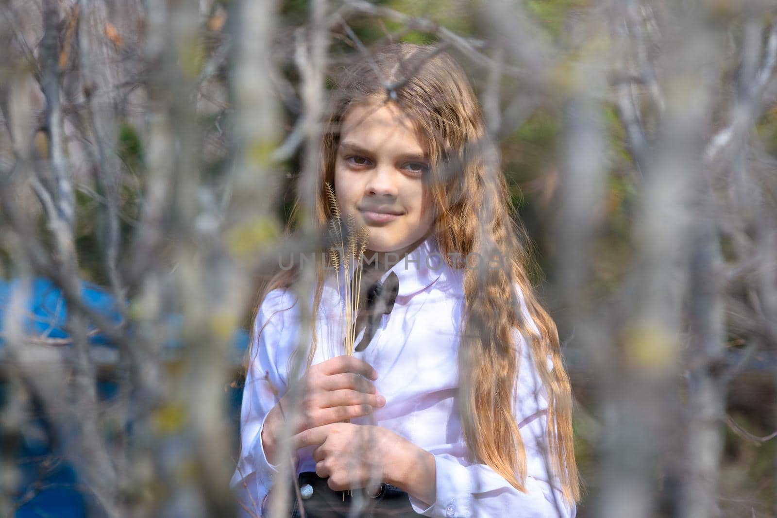 A girl stands with dried wildflowers, in the foreground blurred branches of bushes