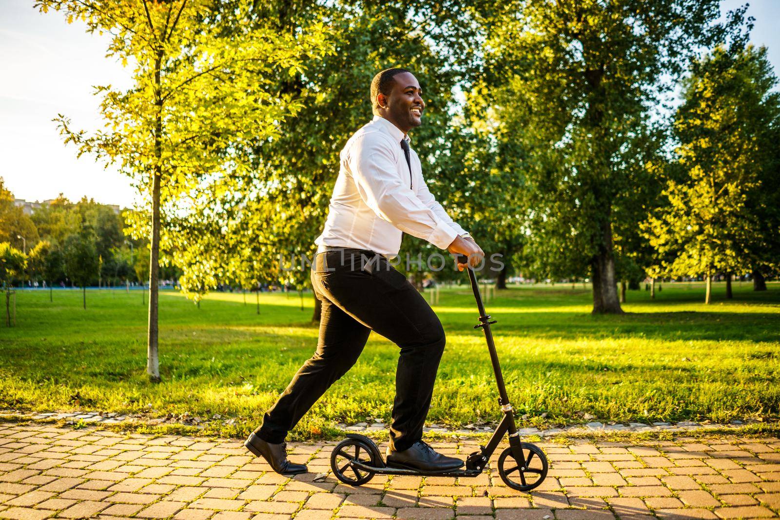 Happy african-american businessman is riding scooter in park on sunny day.