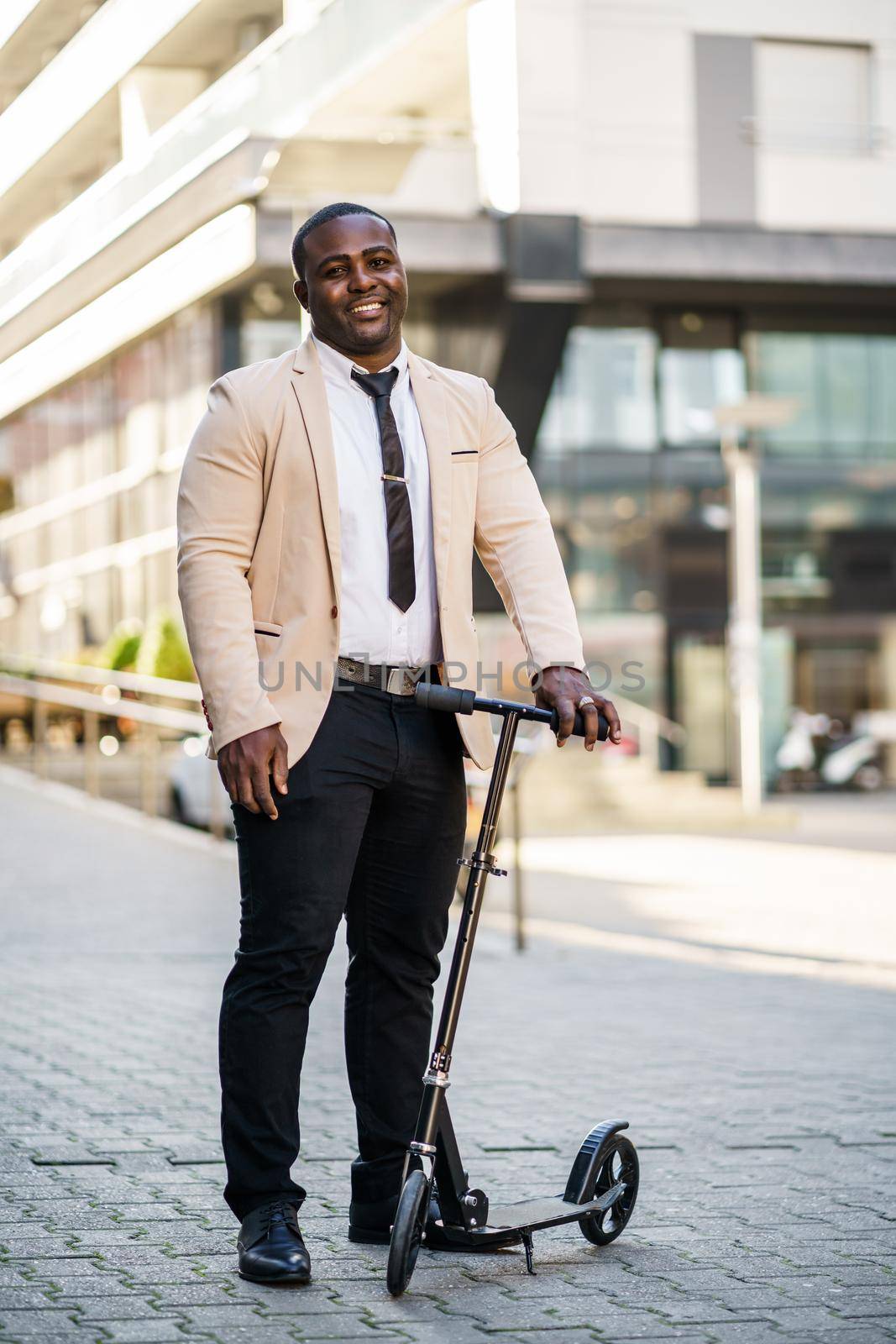 Happy african-american businessman is standing with scooter in front of company building.