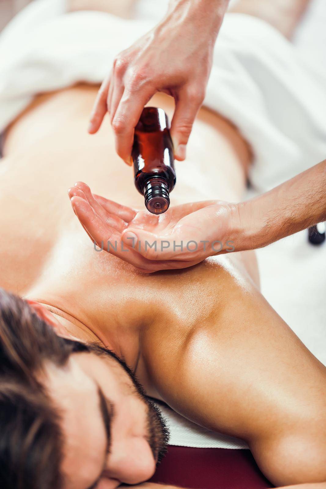 Young man is enjoying massage on spa treatment. Professional masseur is applying massage oil to his skin.