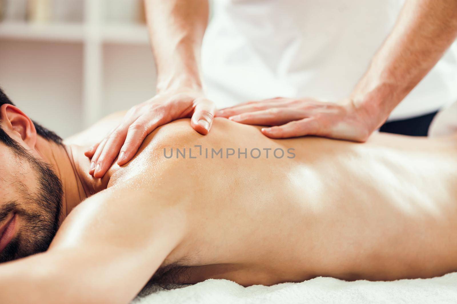 Young man is enjoying massage on spa treatment. Professional masseur is massaging his back.