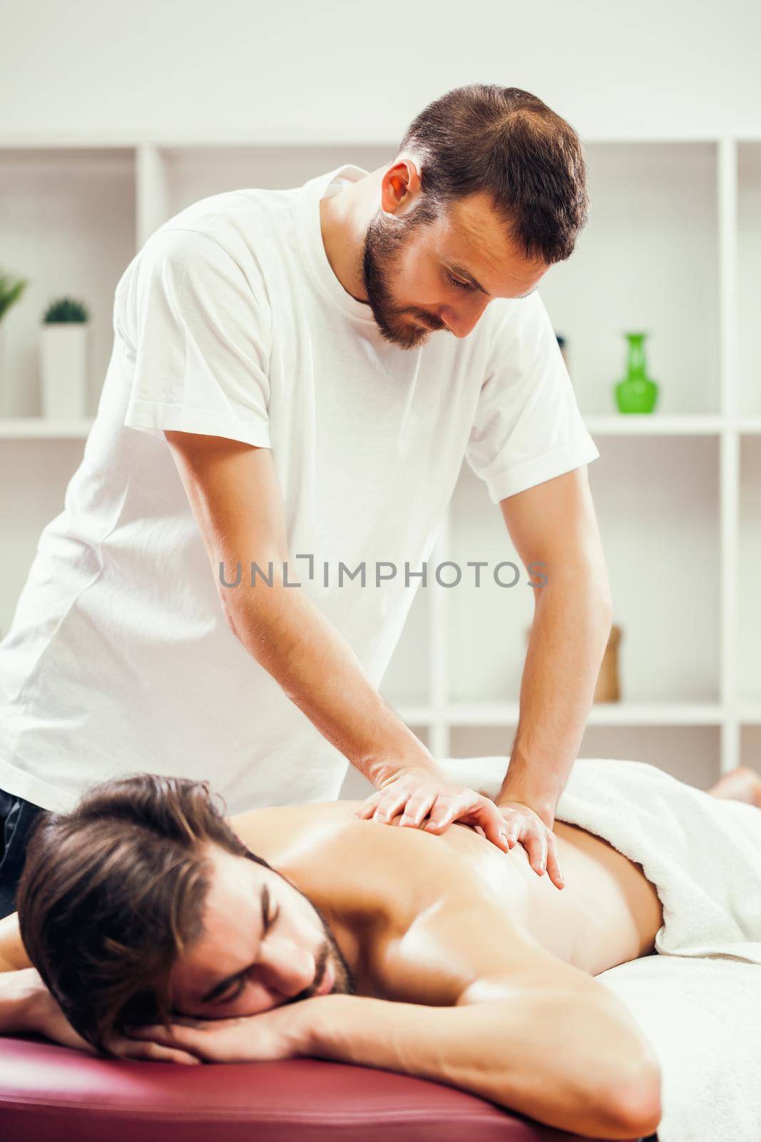 Young man is enjoying massage on spa treatment. Professional masseur is massaging his back.